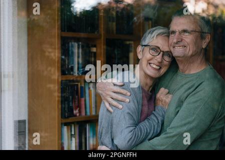 Portrait of happy senior couple hugging behind windowpane Stock Photo