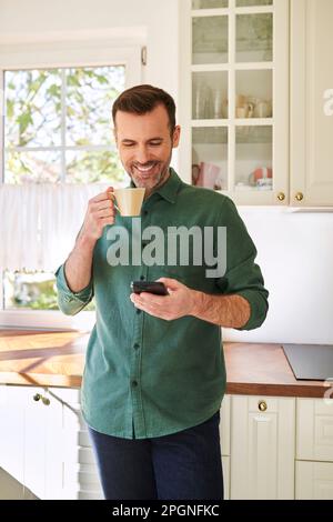 Smiling man using phone while drinking coffee in his kitchen Stock Photo