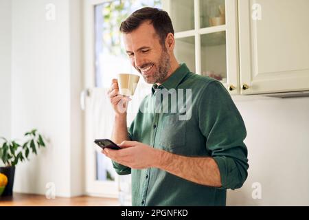 Smiling man using phone while drinking coffee in his kitchen Stock Photo