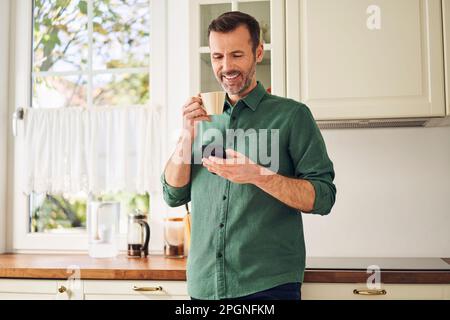 Smiling man using phone while drinking coffee in his kitchen Stock Photo