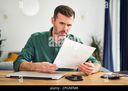 Upset man looking at document with financial figuers while sitting at table at home Stock Photo