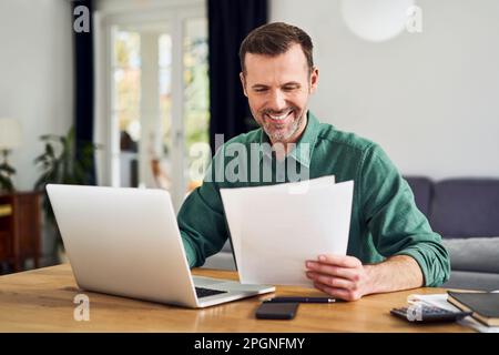 Smiling man working from home using laptop holding documents Stock Photo