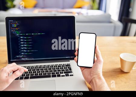 Man working on laptop at home holding phone with blank screen Stock Photo
