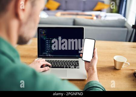 Man working on laptop at home holding phone with blank screen Stock Photo