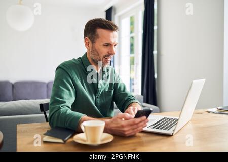 Confident man at home sitting at table using laptop paying with mobile phone Stock Photo