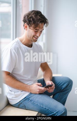 Happy young man using smart phone by window Stock Photo