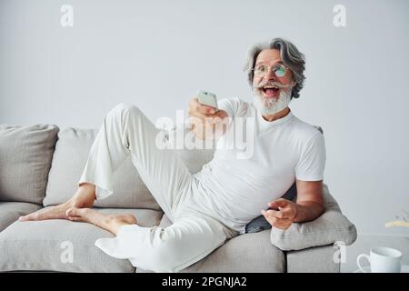Sits on the sofa. Senior stylish modern man with grey hair and beard indoors Stock Photo