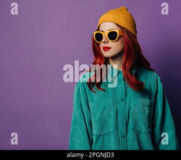 Redhead woman wearing knit hat and sunglasses standing against purple background Stock Photo