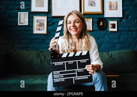Happy actress holding clapboard sitting on sofa at film set Stock Photo