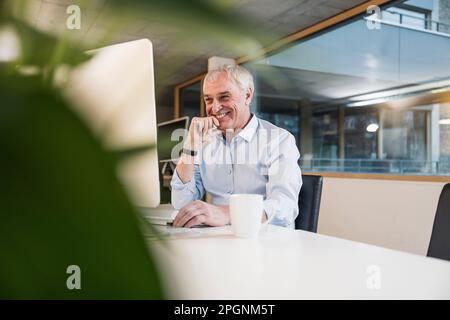 Smiling elderly businessman with hand on chin using computer at office Stock Photo
