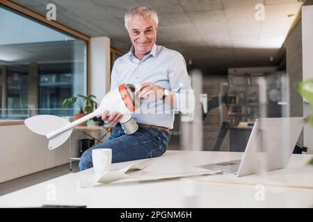 Smiling senior engineer holding wind turbine rotor sitting on desk at office Stock Photo