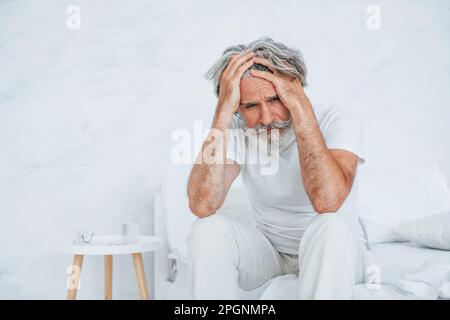 Feels sick. On the bed. Senior stylish modern man with grey hair and beard indoors Stock Photo