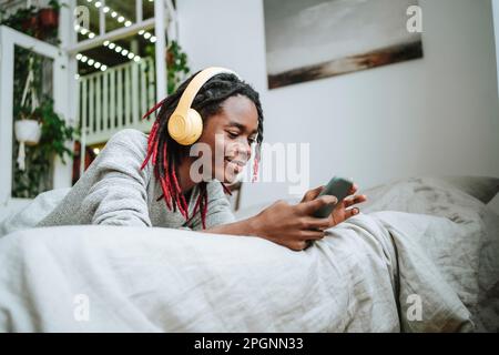 Smiling young man text messaging through smart phone lying on bed at home Stock Photo