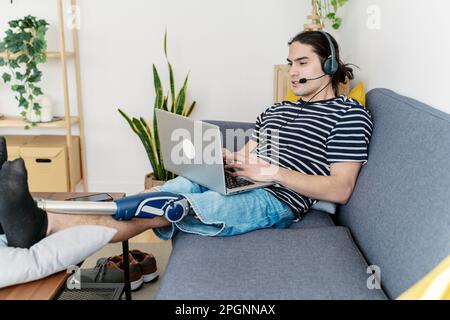 Freelancer with prosthetic leg using laptop wearing headset sitting on sofa at home Stock Photo