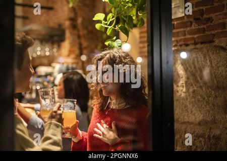Young woman with glass of beer standing with friend seen through window Stock Photo