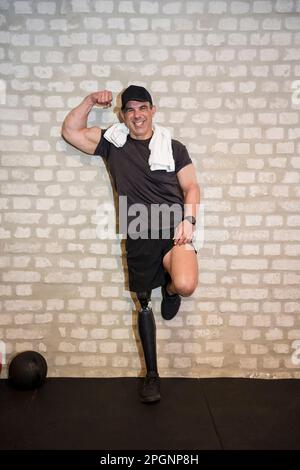 Happy man with prosthetic leg flexing muscle near wall in gym Stock Photo