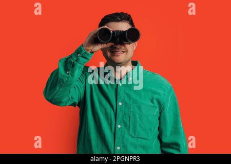 Happy man looking through binoculars standing against coral background Stock Photo