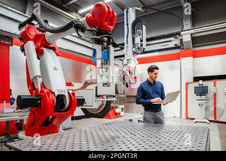 Engineer using laptop standing in robot factory Stock Photo