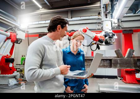 Smiling engineer standing with colleague holding laptop in robot factory Stock Photo
