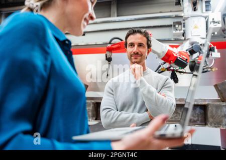 Smiling engineer with hand on chin looking at colleague in robot factory Stock Photo