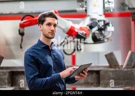 Contemplative engineer holding tablet PC in front of robotic arm Stock Photo