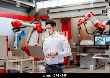 Engineer using laptop standing in robot factory Stock Photo