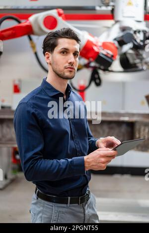Thoughtful engineer with tablet PC standing in robot factory Stock Photo