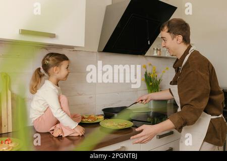 Father making pancakes for daughter sitting on kitchen counter at home Stock Photo