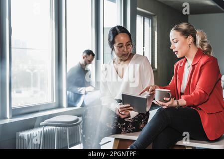 Businesswoman pointing at tablet PC held by colleague sitting on desk in office Stock Photo
