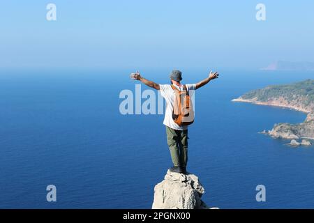 Hiker with arms outstretched standing on edge of cliff looking at sea Stock Photo