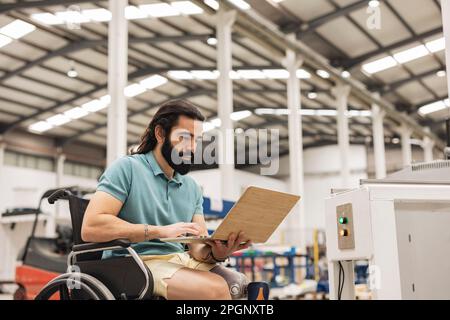 Engineer sitting in wheelchair working on laptop in robotics factory Stock Photo