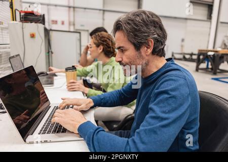 Engineer working on laptop sitting with colleagues in factory Stock Photo