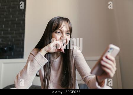 Happy young woman looking at smart phone sitting in bedroom Stock Photo