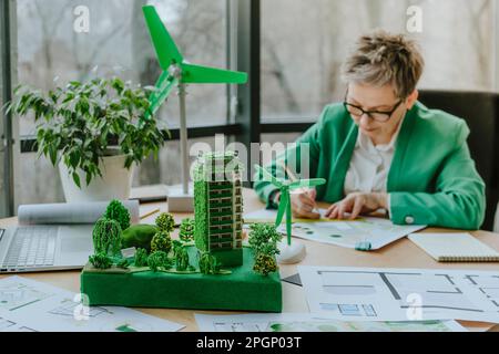 Architect working on blueprint by building and wind turbine models at table in office Stock Photo