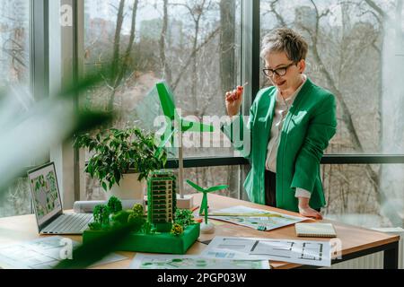 Businesswoman looking at blueprints and biophilic architecture models on desk Stock Photo