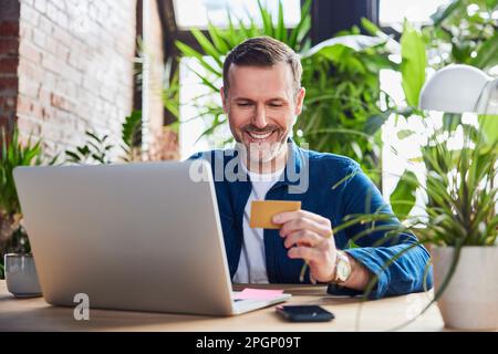 Smiling businessman with laptop holding credit card at loft office Stock Photo