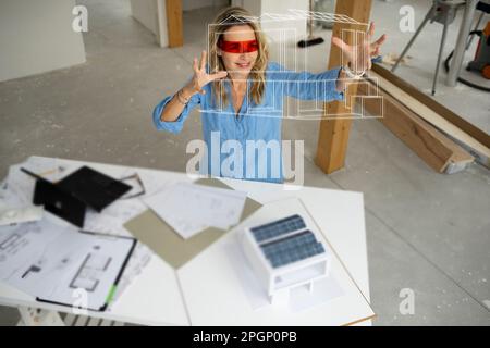 Smiling architect wearing virtual reality simulator gesturing and examining house model Stock Photo