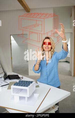 Smiling mature architect wearing virtual reality simulator gesturing and examining house model Stock Photo