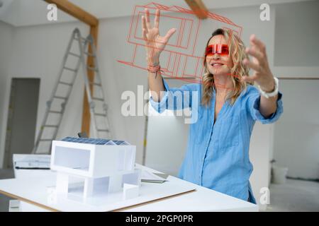 Mature architect wearing virtual reality simulator gesturing and examining house model at site Stock Photo