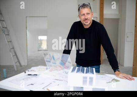 Thoughtful architect with futuristic tablet PC and model home standing at desk Stock Photo