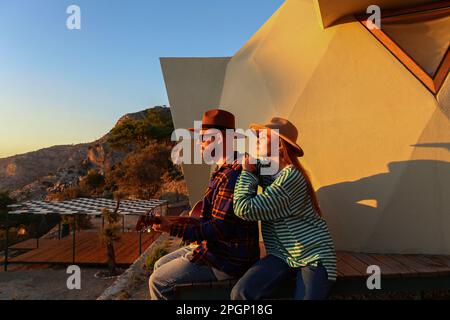Man playing guitar sitting with girlfriend near eco home at sunset Stock Photo