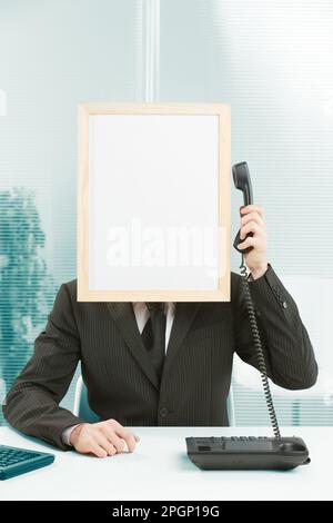 Man with framed sign head in an office answers phone. He has his arm raised with the handset in his hand, which he pulls over to the sign-head to list Stock Photo