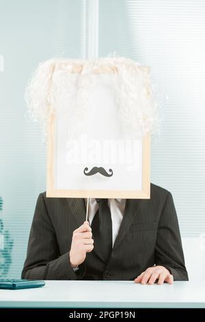 Man with framed sign head in an office. The sign-face also has a white wig and fake mustache added and is ready for you with COPYSPACE. Stock Photo