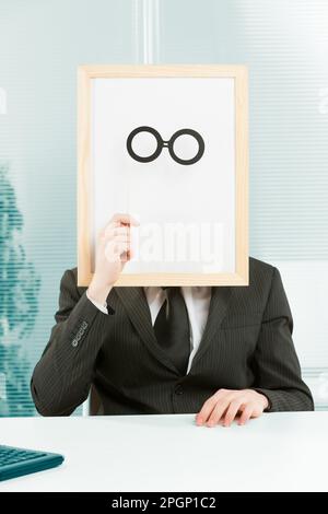 Man with framed sign head in an office. In front of the fake head made of signboard he also places cardboard glasses to complete a disguise that YOU c Stock Photo