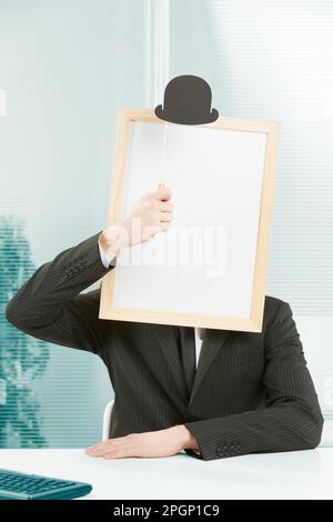 Man with framed sign head in an office. On top of the fake head made of signboard he also puts a bowler hat for a strange and funny but also elegant l Stock Photo