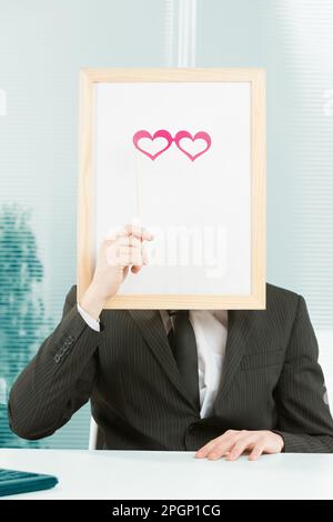 Man with framed sign head in an office. In front of the fake head made of signboard he also puts cardboard glasses in the shape of pink hearts to comp Stock Photo