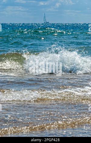 wallpaper with waves - splashing waves in the sun, sailboat on the horizon, blue sky above the horizon, nautical postcard, vertical wallpaper Stock Photo