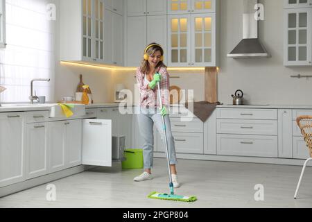 Beautiful young woman with headphones singing while cleaning kitchen Stock Photo