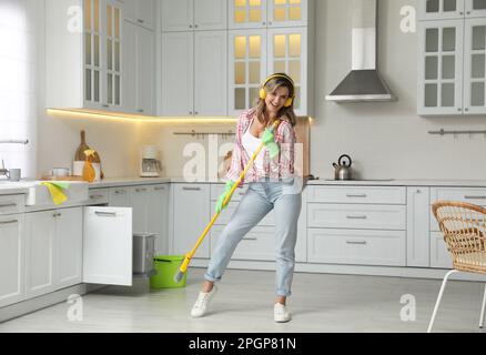 Beautiful young woman with headphones singing while cleaning kitchen Stock Photo