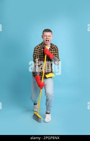 Handsome young man with floor brush singing on light blue background Stock Photo